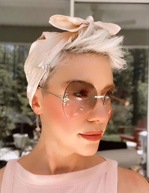Short platinum pixie hair styled with a satin scarf tied into a bow on top, paired with oversized tinted sunglasses.
