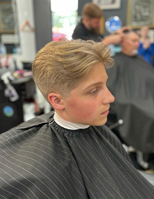 Side-profile of a young male with a side-swept flow haircut, featuring a longer textured top brushed back and a clean low taper on the sides and back.