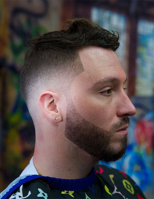 Side profile of a man with a skin fade haircut blended into a sharply lined beard, featuring a clean line-up and short textured hair on top.