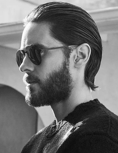 Man with medium-length slicked back hair wearing sunglasses, featuring a full beard and a sleek, polished hairstyle styled smoothly away from the face.