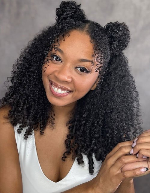 Front view of a woman with natural curly hair styled in half-up space buns, featuring two small buns on top with loose curls left down.