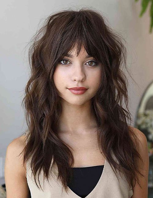 Woman with long wavy hair and bottleneck bangs softly framing her eyes.