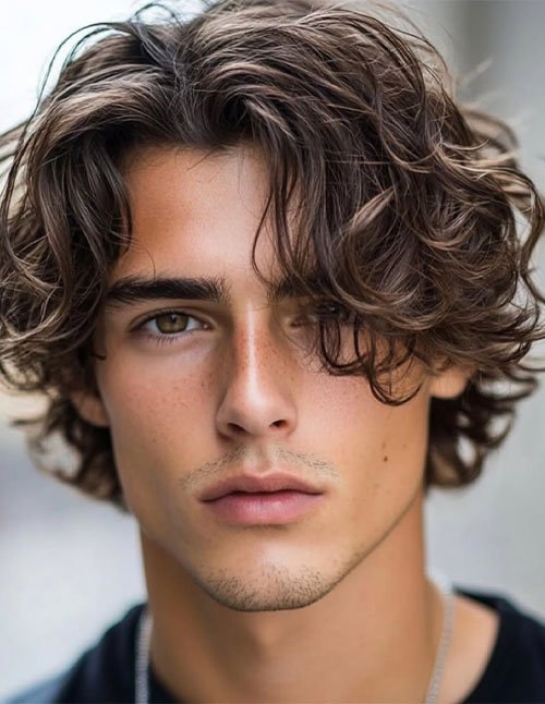 Close-up of a man with medium-length surfer hair, loose curls, and layered texture, with a messy fringe falling toward the eyes.