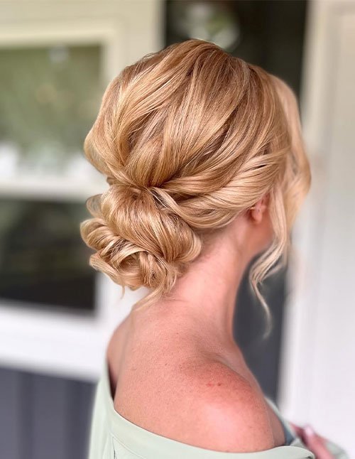 Side view of a woman with blonde hair styled in a textured low updo with soft curled pieces and a loose face-framing strand.