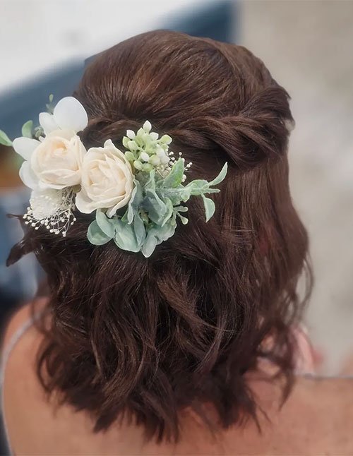 Back view of a twisted half-up hairstyle on lob-length wavy hair, decorated with a white rose and greenery hair piece on one side.
