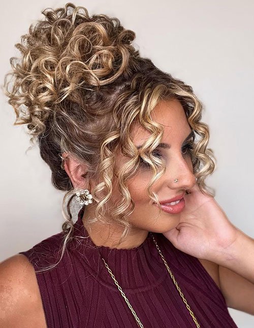 Woman with a curly updo featuring a high textured bun and face-framing spiral curls, shown in side profile.